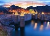Hoover Dam at sunset with illuminated towers, calm water below, surrounding rocky landscape, and a bridge in the background.