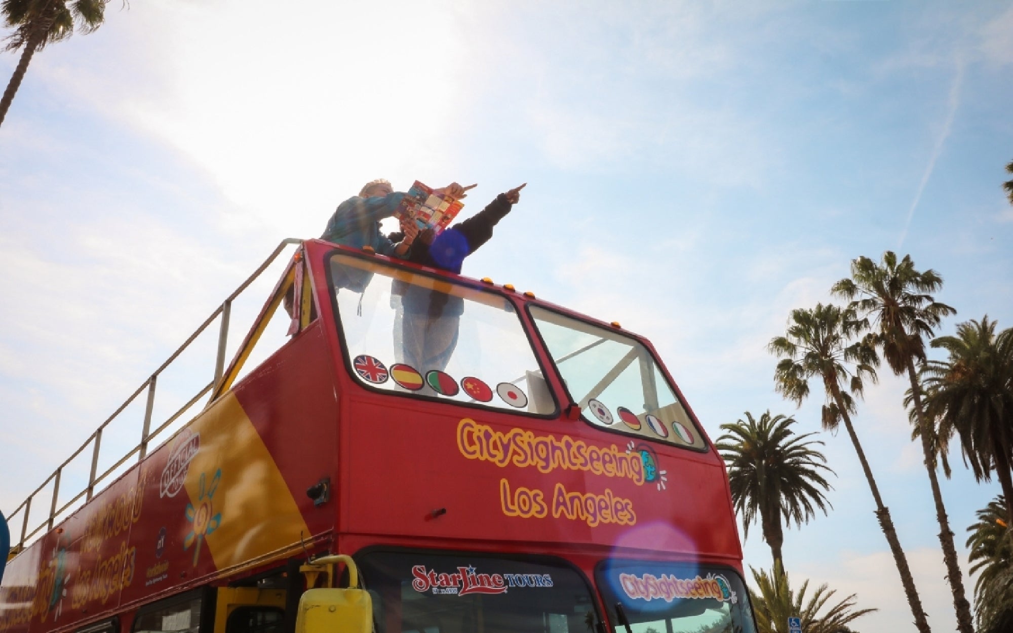 Two people stand on the upper deck of a red CitySightseeing Los Angeles tour bus, holding a map and pointing, with palm trees and a sunny sky in the background.