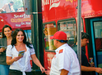 A woman holding a brochure smiles as she walks past a man in a red-and-white uniform near a red double-decker sightseeing bus. Other people are boarding the bus.