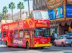 A red double-decker sightseeing bus drives down a street with palm trees and a theater marquee in the background.
