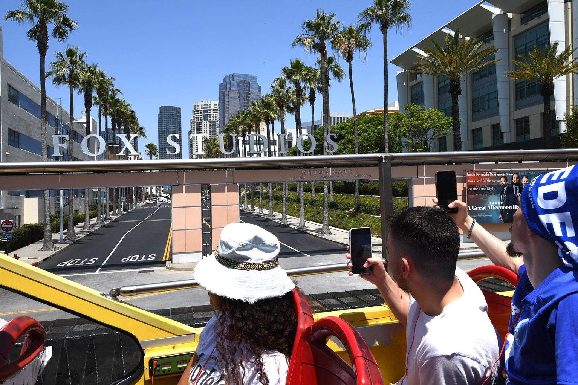 Tourists on a bus take photos of the entrance to Fox Studios, framed by palm trees and city buildings.