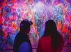 Two people stand in front of a vibrant, multicolored tropical leaf mural.
