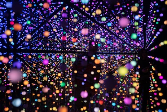 A colorful installation of suspended, glowing lights creates a kaleidoscope effect in a dark room, with shadowy figures in the background.