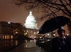 A person holding an umbrella stands in front of the illuminated U.S. Capitol building at night on a rainy evening.