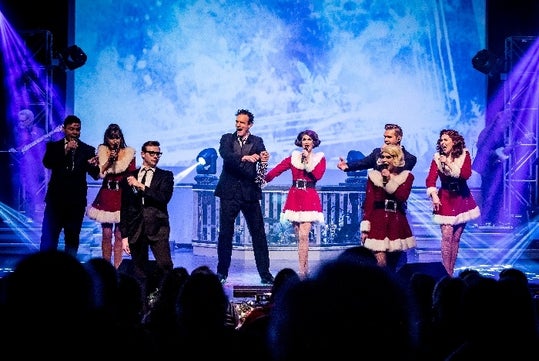 A group of performers sing on stage in festive Christmas outfits, with a blue-lit background and stage lighting, in front of an audience.