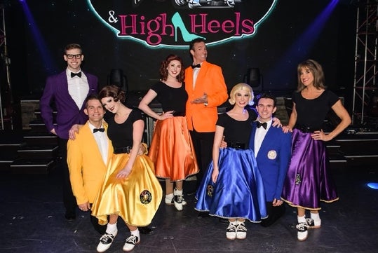 A group of four men in colored tuxedos and four women in matching poodle skirts pose together on stage in front of a “& High Heels” sign.