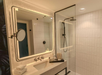 Private bathroom with a large illuminated mirror, wall-mounted sink, glass shower enclosure, rainfall showerhead, and neatly arranged toiletries on the counter.