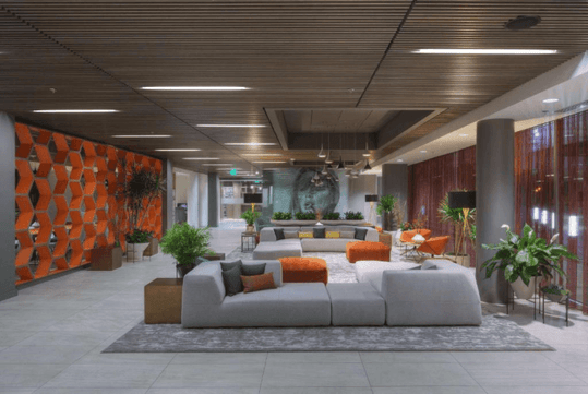 A lobby decorated in gray and orange with several cushioned seating options and large house plants with a mural of a woman on the far end of the room.