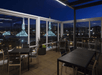 A rooftop patio with glass walls and no roof, several black square tables with matching chairs, and a great view of the city of Portland at night.