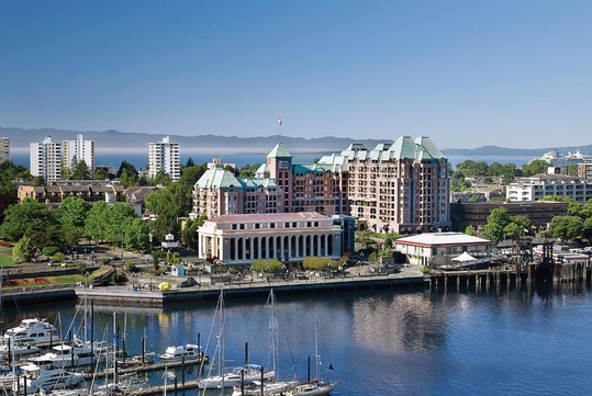 Aerial view of a waterfront cityscape with boats docked in the foreground, surrounded by mid-rise buildings and greenery under a clear blue sky.