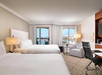 Hotel room with two double beds, a large window with city view, a seating area with a gray chair, a desk with black chair, and neutral decor.