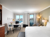 Hotel room with a bed, work desk, chair, and a view of water and buildings through large windows. The room has neutral decor and a modern style.