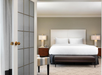 Modern hotel room with a neatly made double bed, two bedside tables with lamps, a bench at the foot of the bed, and an open door with glass panels.