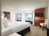 Hotel room with a large bed, wall-mounted TV, wooden furniture, patterned carpet, striped chair, and a window view of water and buildings.