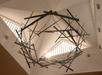 Sculptural metal chandelier with intersecting rods hangs from a high ceiling with triangular skylights, sunlight streaming through the windows.