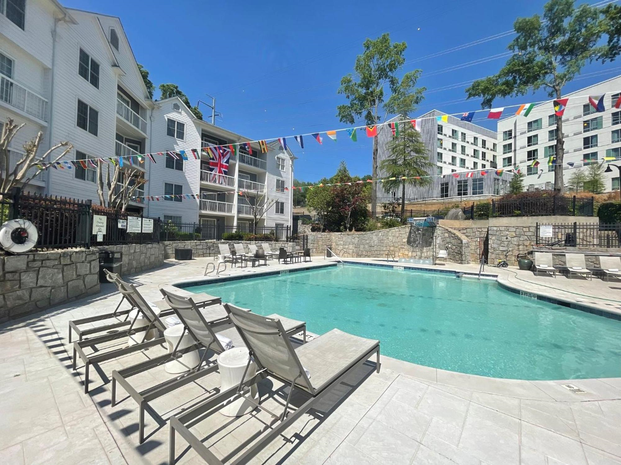 Outdoor swimming pool with lounge chairs, surrounded by apartment buildings and stone walls; string of international flags hung above the pool; clear, sunny day.