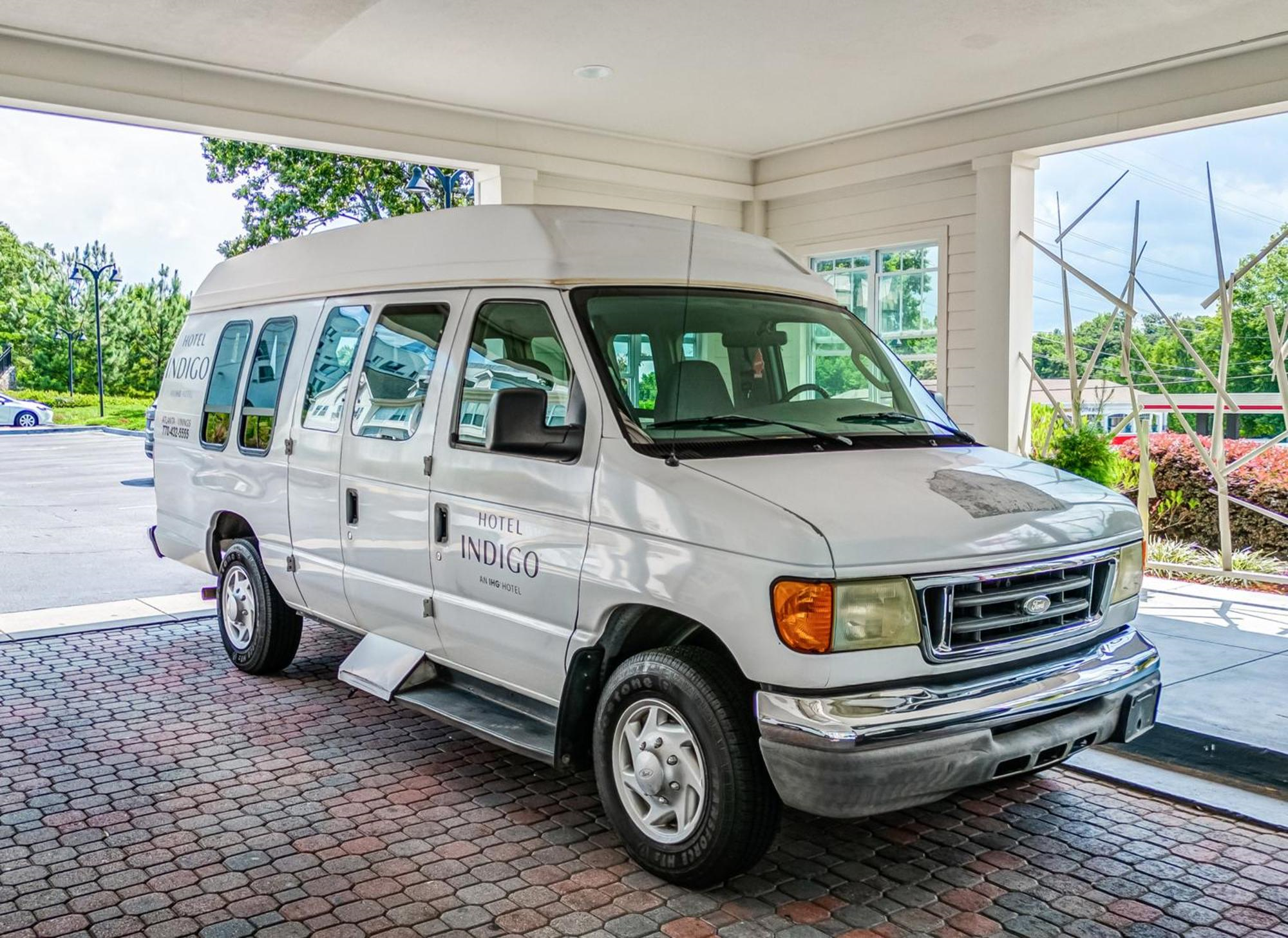White Hotel Indigo van parked under a covered driveway with a side step extended, surrounded by landscaped greenery and brick pavement.