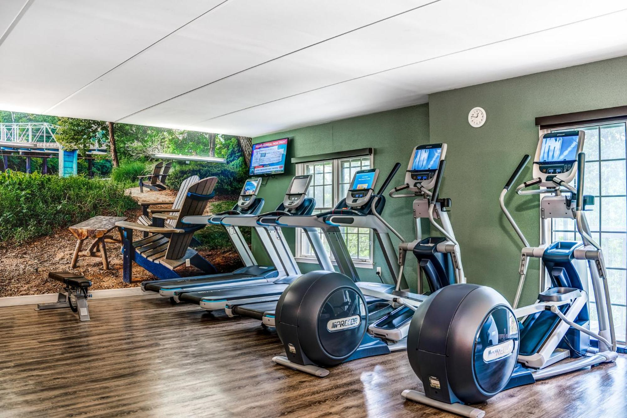 A fitness room with four cardio machines—two treadmills and two elliptical trainers—facing wall-mounted TVs, next to large windows and a wall mural of an outdoor scene.
