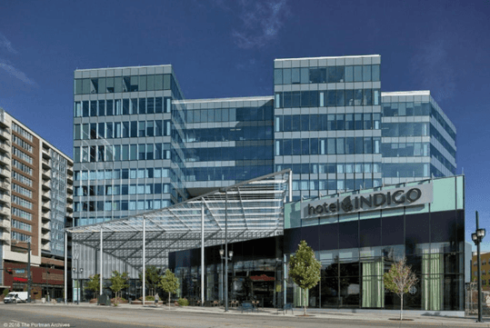 The blue glass front exterior of the Hotel Indigo Denver Downtown with small trees lining the walkway on a bright and sunny day