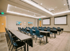 A meeting room with rows of gray tables with blue chairs lined up in front of two large projector screens with art on the far wall.