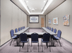 A meeting room with long gray tables in a "U" formation with blue chairs lining the outside with a projector screen at the far end of the room.