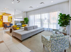 Modern lobby with gray sofas, patterned chairs, and large plants. A yellow-accented bookshelf and fireplace are in the background, with large windows allowing natural light.