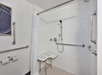 Accessible shower with white tiles, a folding seat, and grab bars. A showerhead is attached on the right wall. Toilet paper holders and a partial curtain are visible on the left.