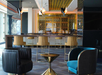 Modern lounge area with blue and black armchairs, a gold-accented bar, and high chairs. Overhead lights illuminate the space, which features reflective surfaces and geometric patterns.