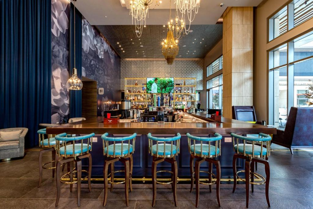 Chic bar interior with a long wooden counter, blue cushioned stools, chandeliers, and a decorative wall mural. Shelves with bottles and glasses in the background.