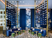 Modern hotel lobby with blue velvet seating, gold accents, patterned carpet, and decorative shelves. Large floral wall art and pendant lights enhance the chic ambiance. Computers are on desks.