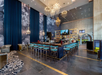 Spacious, modern bar with high ceilings, chandeliers, and a long, blue bar counter surrounded by chairs. Floral wallpaper and two TV screens adorn the walls.