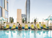 Two people by a rooftop pool with yellow and blue lounge chairs, striped umbrellas, and tall buildings in the background on a clear day.