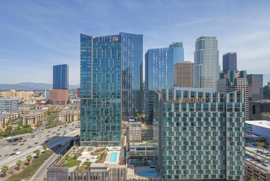 Aerial view of skyscrapers in downtown Los Angeles, featuring glass facades, a rooftop pool, and bustling streets below under a clear blue sky.