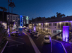 A well-lit motel parking lot at night with parked cars, surrounded by palm trees; a multi-story hotel is visible in the background.