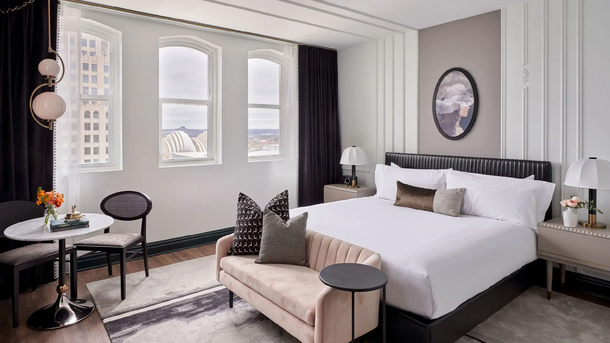 Modern hotel room with a large bed, two nightstands, a small round table with two chairs, decorative pillows, and tall windows with city views.