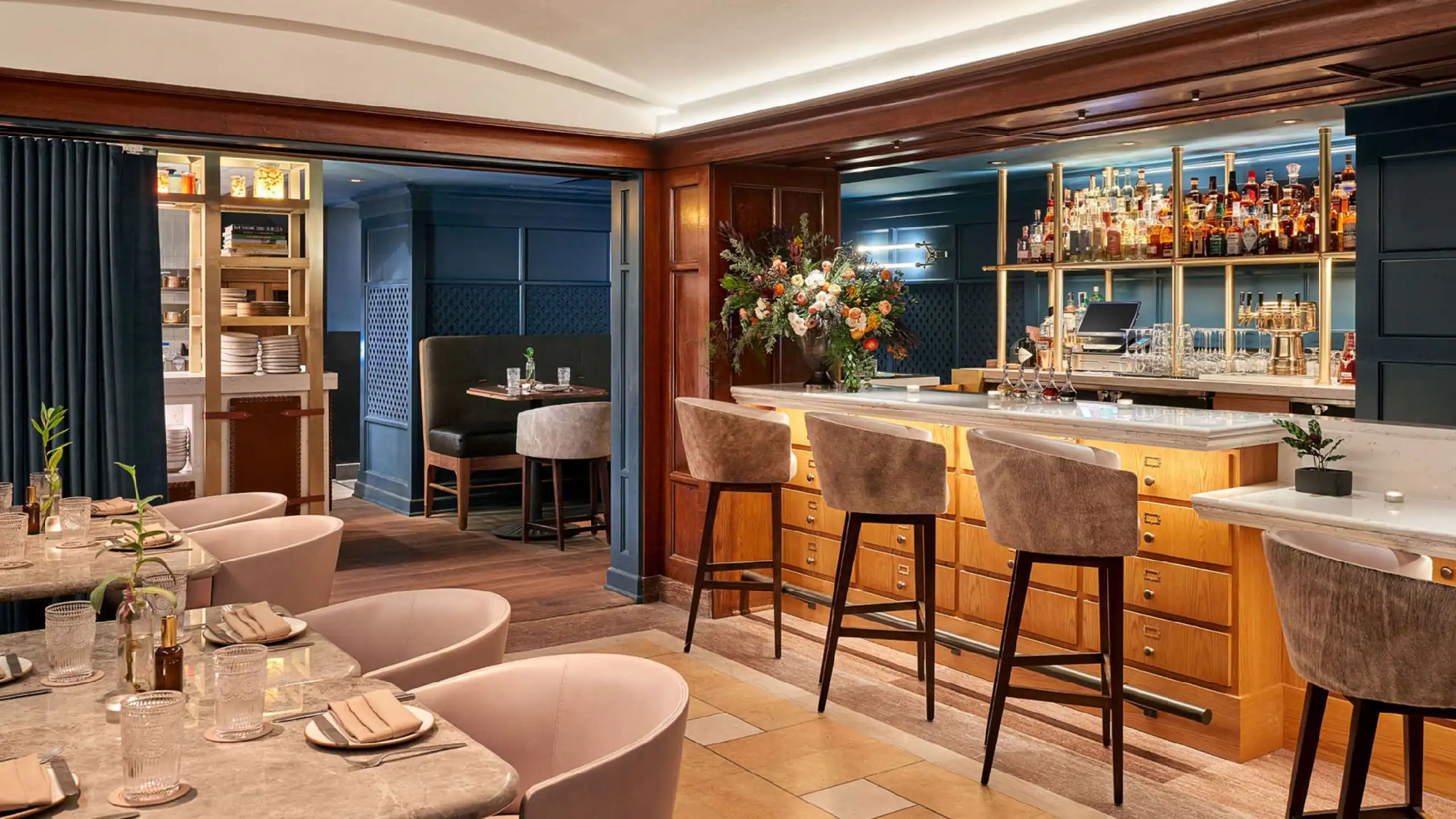 Modern restaurant interior with a marble bar, high chairs, dining tables, and a backlit liquor display; floral arrangement and booth seating in the background.