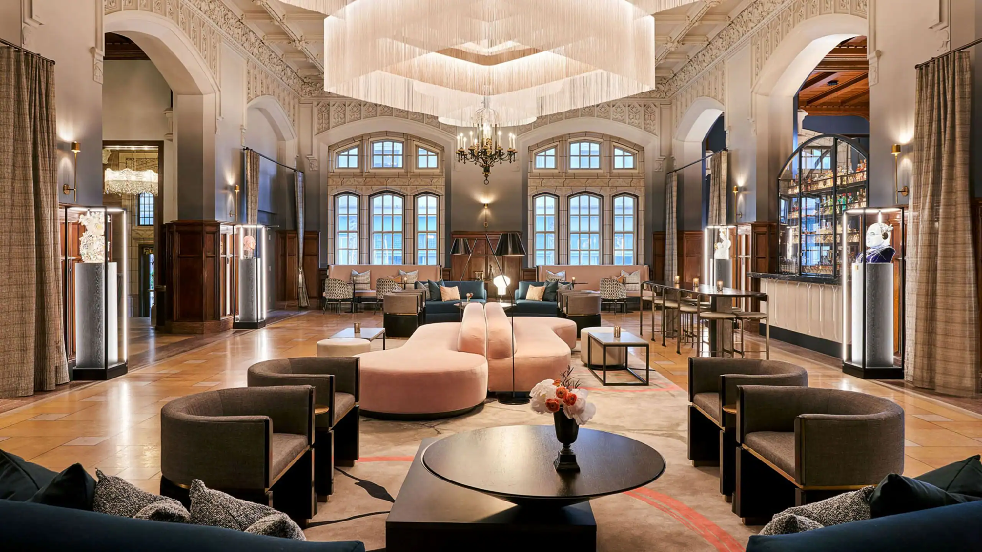 Spacious hotel lobby with high ceilings, large windows, modern chandeliers, pink and gray seating, and a central coffee table with a vase of flowers.
