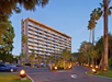 A multi-story hotel with balconies is surrounded by palm trees. The entrance features a circular driveway with decorative lights and parked cars.