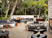 Stylish outdoor patio with curved seating, tables, and decorative cushions surrounded by lush greenery and trees. A bar area is visible with various drinks and decor.