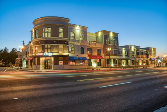 Street view of a multi-story building at dusk with illuminated windows and storefronts. Light trails from passing vehicles are visible on the road.