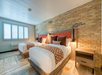 A hotel room with two double beds, orange and white bedding, a wooden nightstand, brick accent wall, and a window with shutters.