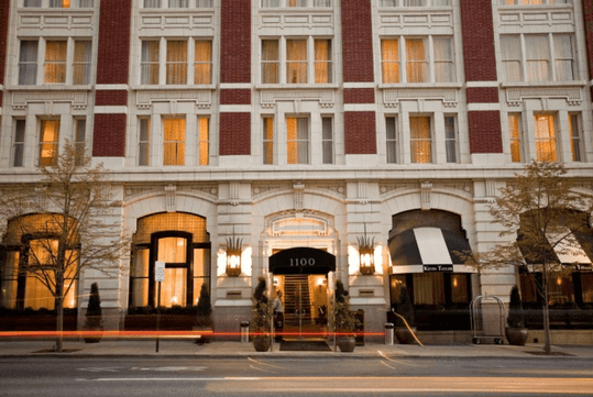 The front entrance of the Hotel Teatro with small trees along the sidewalk and the rows of windows glowing with light/