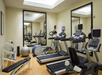 A small fitness center with a row of cardio equipment and a small free weights section with a bench and to mirrors on the walls.