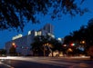 A modern building illuminated at night with light trails from passing cars at an intersection, surrounded by trees and city structures.