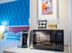 A hotel room counter with a microwave, a coffee maker, tea bags, and cups in front of a bed and a colorful framed artwork on the wall.