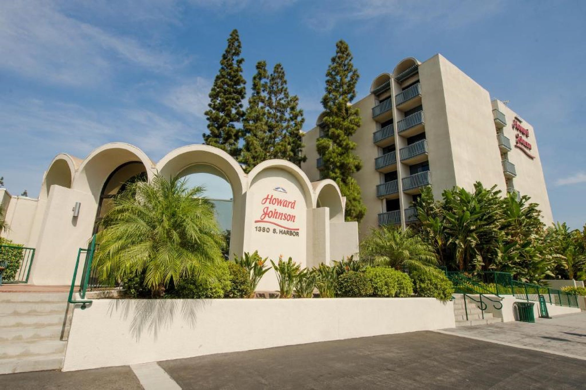 Howard Johnson hotel building with arched entrance, surrounding greenery, and a multi-story section with balconies under a clear blue sky.