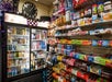 Shelves stocked with snacks and protein bars next to a refrigerated display of drinks in a convenience store decorated with Halloween garland and a “BOO” sign.
