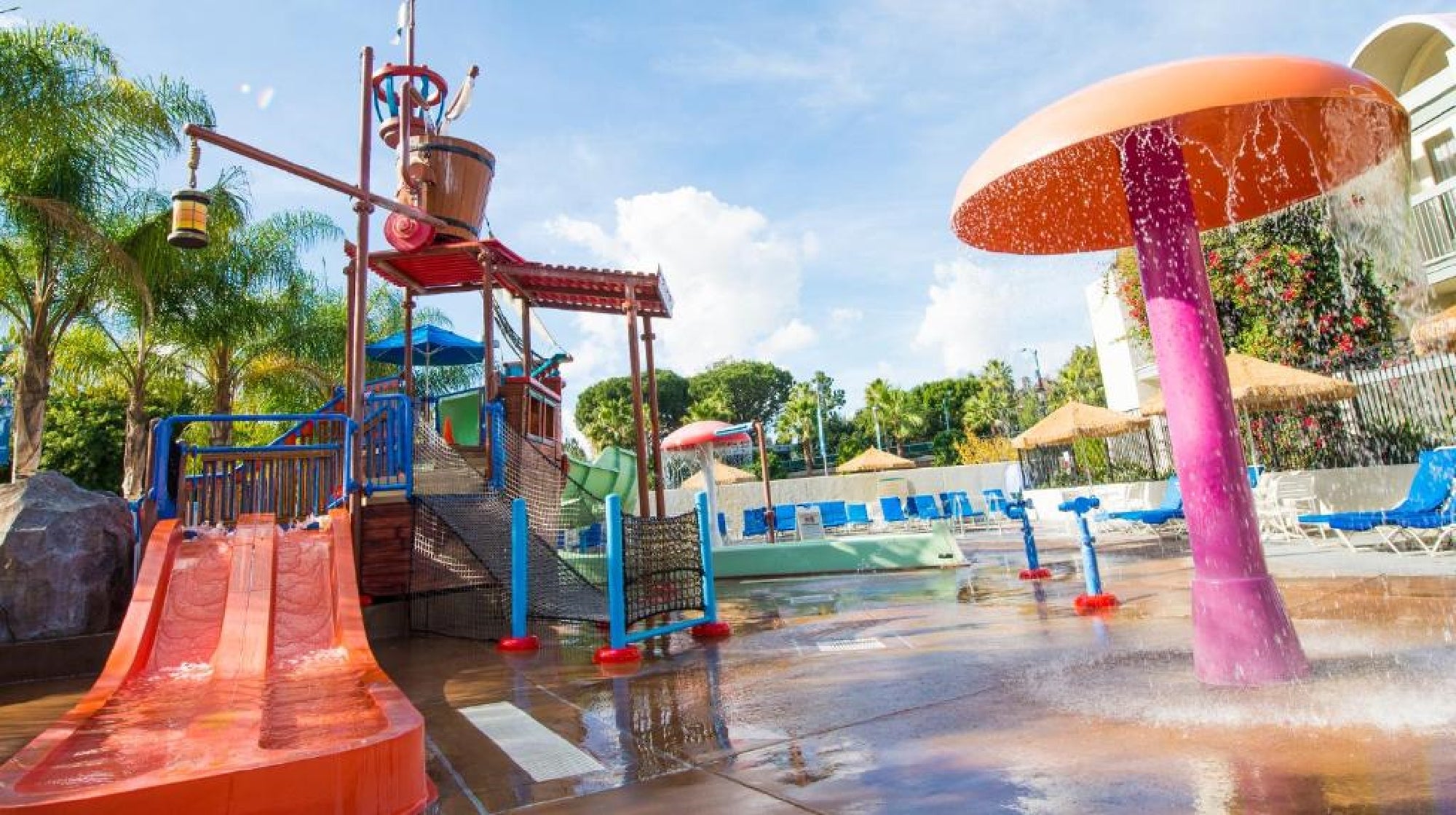 Outdoor children's water play area with slides, a large tipping bucket, sprayers, and a mushroom-shaped fountain, surrounded by lounge chairs and palm trees.