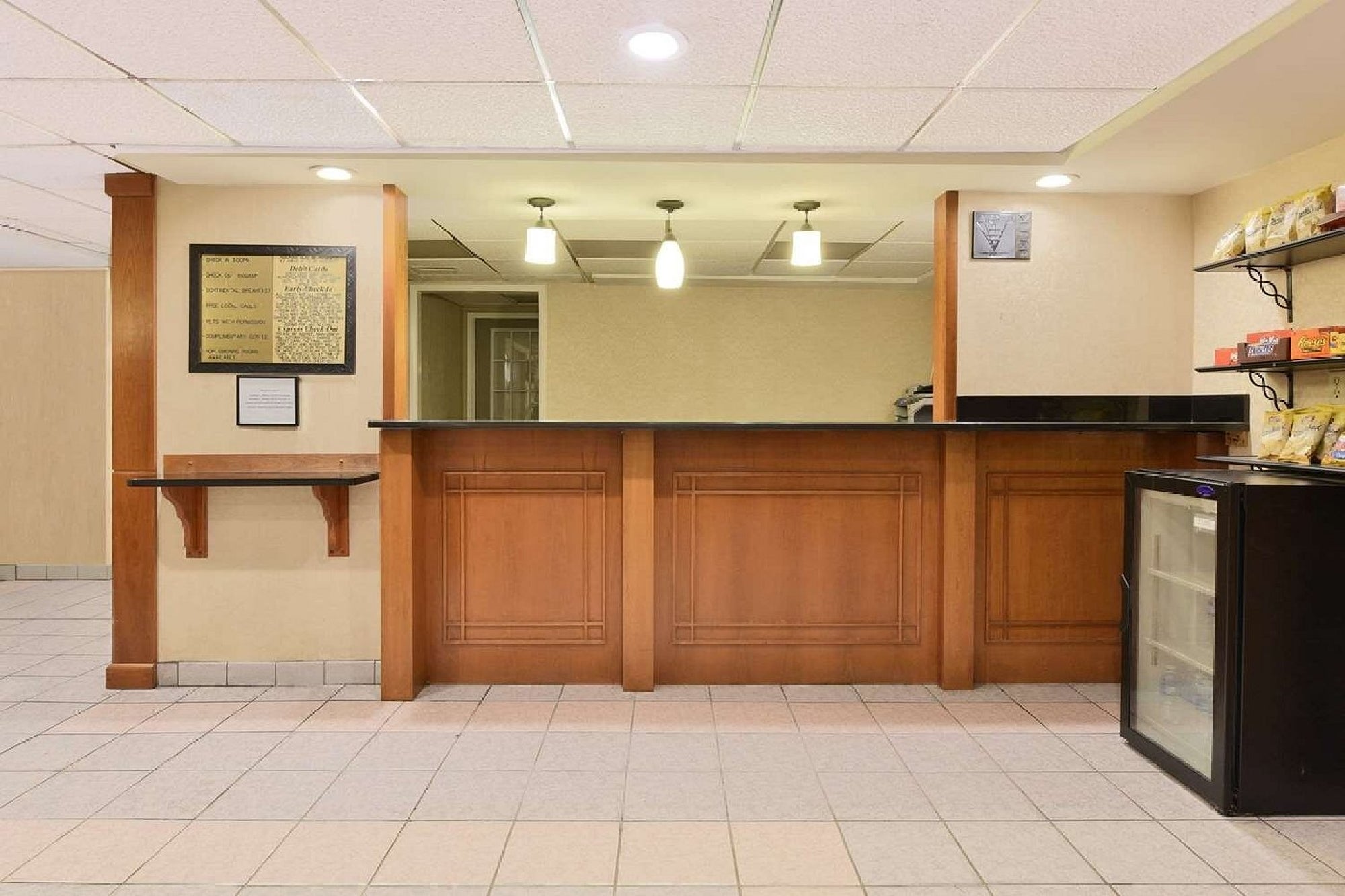 A hotel reception desk with wooden paneling, three pendant lights overhead, a small fridge with glass door on the right, and plaques mounted on the wall to the left.