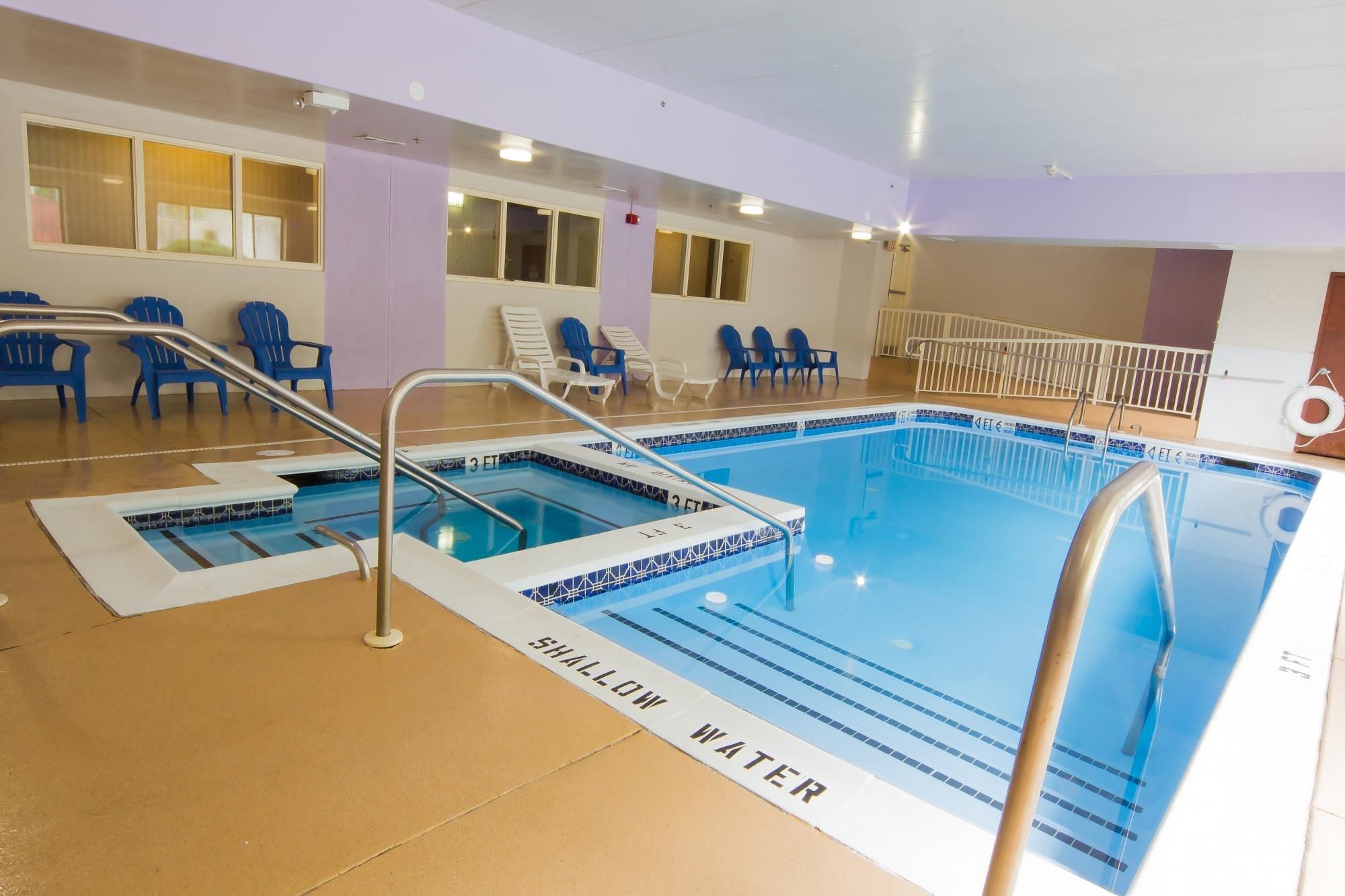 Indoor swimming pool with a hot tub, surrounded by lounge chairs and blue chairs; labeled "shallow water" area and metal handrails for entering the pool.
